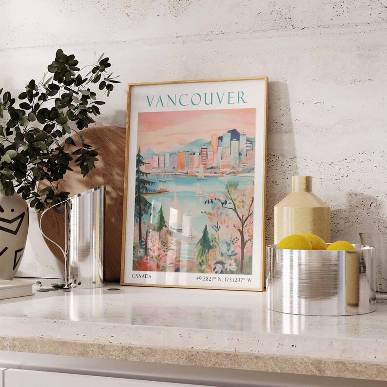 May include: A vintage-style poster with a gold frame depicting a cityscape of Vancouver, Canada. The poster has a pink and blue color scheme and features the text "Vancouver" and "Canada" along with the coordinates "49.2827&deg; N, 123.1207&deg; W".