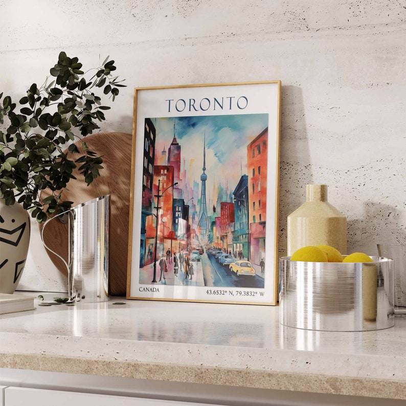Op de afbeelding: Een vintage poster met een kleurrijke illustratie van een straatbeeld in Toronto, Canada. De poster heeft een gouden lijst en de tekst "TORONTO" in witte letters bovenaan. De tekst "CANADA" staat linksonder op de poster. De poster staat op een aanrecht met een zilveren kan, een houten snijplank, een vaas met een plant en een zilveren kom met citroenen.