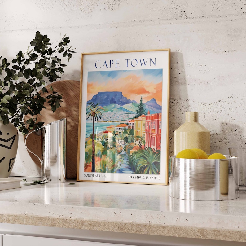 May include: A vintage-style poster with a gold frame depicting a colorful illustration of Cape Town, South Africa. The poster includes the text "CAPE TOWN", "SOUTH AFRICA", and "33.9249&deg; S, 18.4241&deg; E".