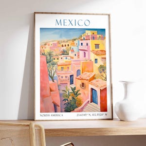 Op de afbeelding: Een kleurrijk aquarel schilderij van een stadsgezicht in Mexico. De gebouwen zijn geschilderd in tinten roze, geel en oranje. Het schilderij is ingelijst in een houten lijst.