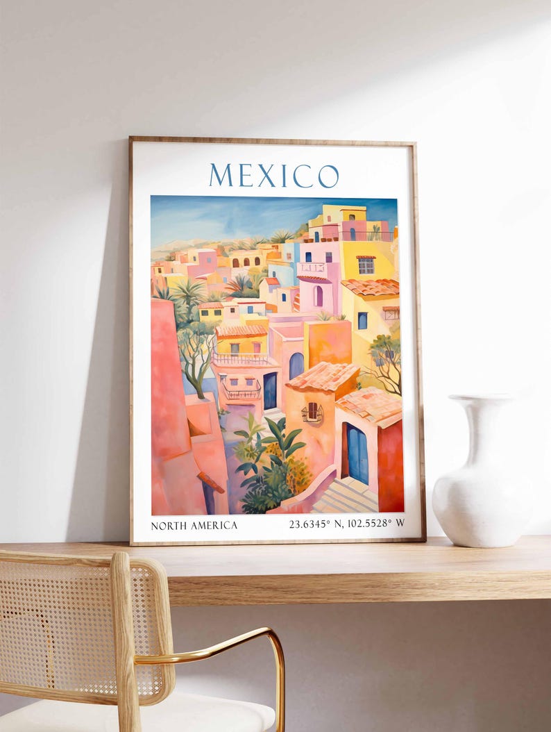 Op de afbeelding: Een kleurrijk aquarel schilderij van een stadsgezicht in Mexico. De gebouwen zijn geschilderd in tinten roze, geel en oranje. Het schilderij is ingelijst in een houten lijst.