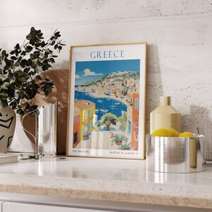 May include: A framed vintage travel poster with a colorful illustration of a Greek island town with a blue bay and the text "GREECE THE BALKANS 39.0742&deg; N, 21.8243&deg; E".