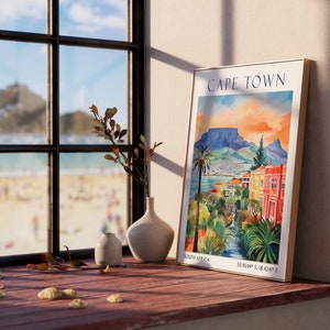 May include: A framed poster with a colorful illustration of Cape Town, South Africa. The poster features a mountain range, a cityscape, and a river. The text "CAPE TOWN" is printed in blue at the top of the poster. The poster is hanging on a wall next to a window with a view of a beach.