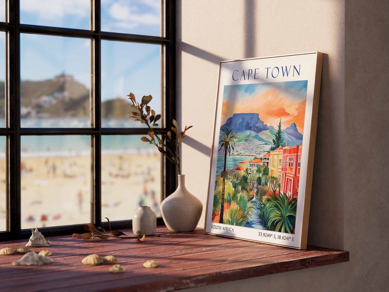 May include: A framed poster with a colorful illustration of Cape Town, South Africa. The poster features a mountain range, a cityscape, and a river. The text "CAPE TOWN" is printed in blue at the top of the poster. The poster is hanging on a wall next to a window with a view of a beach.