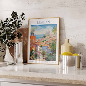 May include: A framed poster with a watercolor illustration of Lisbon, Portugal. The poster has a gold frame and features a cityscape with colorful buildings and a blue ocean in the background. The text "LISBON" is printed in large, bold letters at the top of the poster. The poster is sitting on a white countertop with a silver pitcher and a silver bowl with lemons.