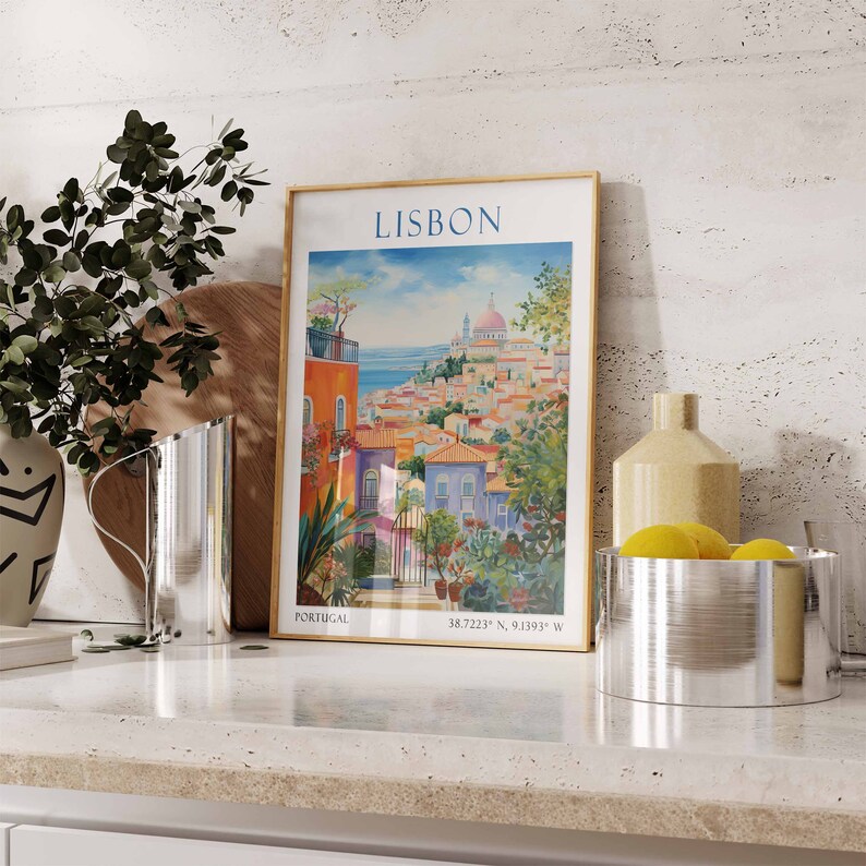 May include: A framed poster with a watercolor illustration of Lisbon, Portugal. The poster has a gold frame and features a cityscape with colorful buildings and a blue ocean in the background. The text "LISBON" is printed in large, bold letters at the top of the poster. The poster is sitting on a white countertop with a silver pitcher and a silver bowl with lemons.