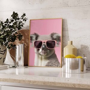 May include: A framed print of a koala wearing pink sunglasses against a pink background. The koala is looking directly at the camera.