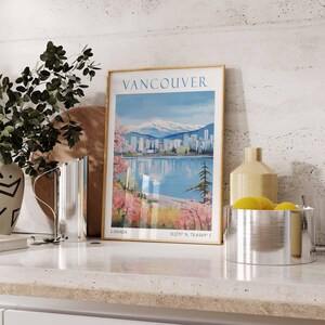 Puede incluir: Un p&oacute;ster vintage enmarcado con una ciudad y monta&ntilde;as en el fondo. El p&oacute;ster se titula "Vancouver" y tiene el texto "Canad&aacute;" y "49.27&deg; N, 78.6569&deg; E" en la parte inferior. El p&oacute;ster est&aacute; sobre una encimera con una jarra plateada, un jarr&oacute;n con limones amarillos y un taz&oacute;n plateado.