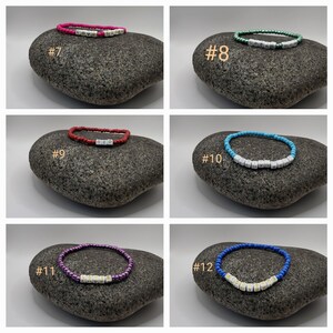 K&ouml;nnte beinhalten: Sechs bunte Perlenarmb&auml;nder mit wei&szlig;en Buchstabenperlen, die verschiedene W&ouml;rter buchstabieren. Die Armb&auml;nder werden auf grauen Steinen pr&auml;sentiert. Die Armb&auml;nder sind von 7 bis 12 nummeriert.