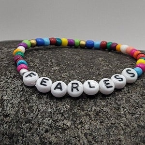 K&ouml;nnte beinhalten: Ein buntes Perlenarmband mit dem Wort "FEARLESS" in wei&szlig;en Perlen mit schwarzen Buchstaben.