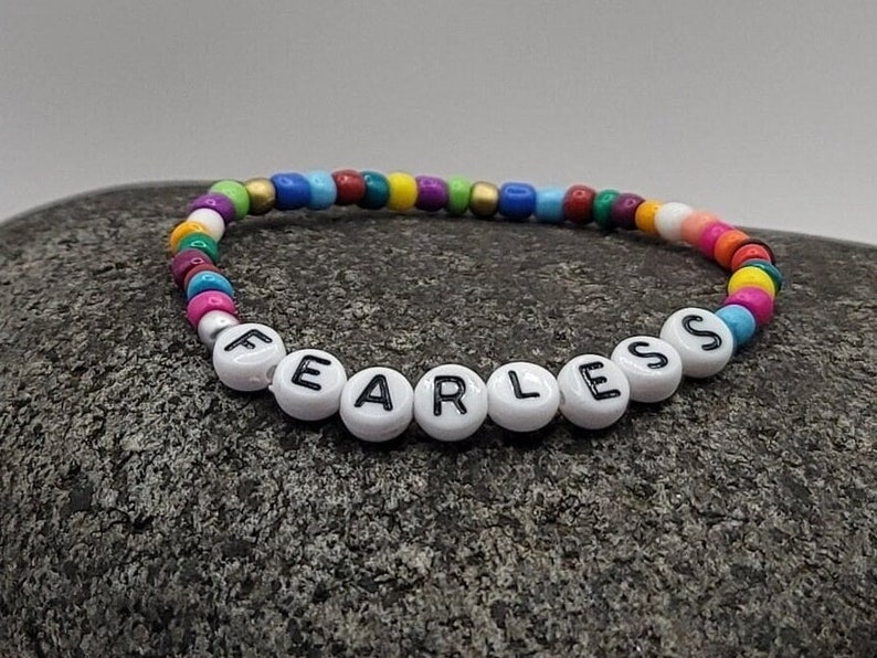K&ouml;nnte beinhalten: Ein buntes Perlenarmband mit dem Wort "FEARLESS" in wei&szlig;en Perlen mit schwarzen Buchstaben.
