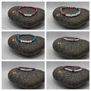 K&ouml;nnte beinhalten: Sechs bunte Perlenarmb&auml;nder mit verschiedenen W&ouml;rtern, die in wei&szlig;en Buchstaben ausgeschrieben sind. Die Armb&auml;nder liegen auf einem grauen Gesteinsuntergrund. Die W&ouml;rter sind "RED", "LOVER", "1985", "LENDERHAZE", "BLESS" und "CRUEL SUN".