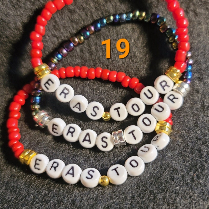 K&ouml;nnte beinhalten: Drei rote Perlenarmb&auml;nder mit wei&szlig;en Perlen, die den Namen "Erasto" in Gro&szlig;buchstaben buchstabieren. Die Armb&auml;nder sind &uuml;bereinander gelegt und die Zahl "19" ist in Orange &uuml;ber den Armb&auml;ndern geschrieben.