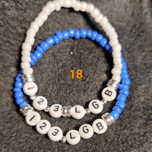 K&ouml;nnte beinhalten: Zwei Perlenarmb&auml;nder mit wei&szlig;en und blauen Perlen. Die Armb&auml;nder haben wei&szlig;e Perlen mit den Buchstaben "123 L G B" darauf. Die Zahl "18" ist auf dem Bild geschrieben.