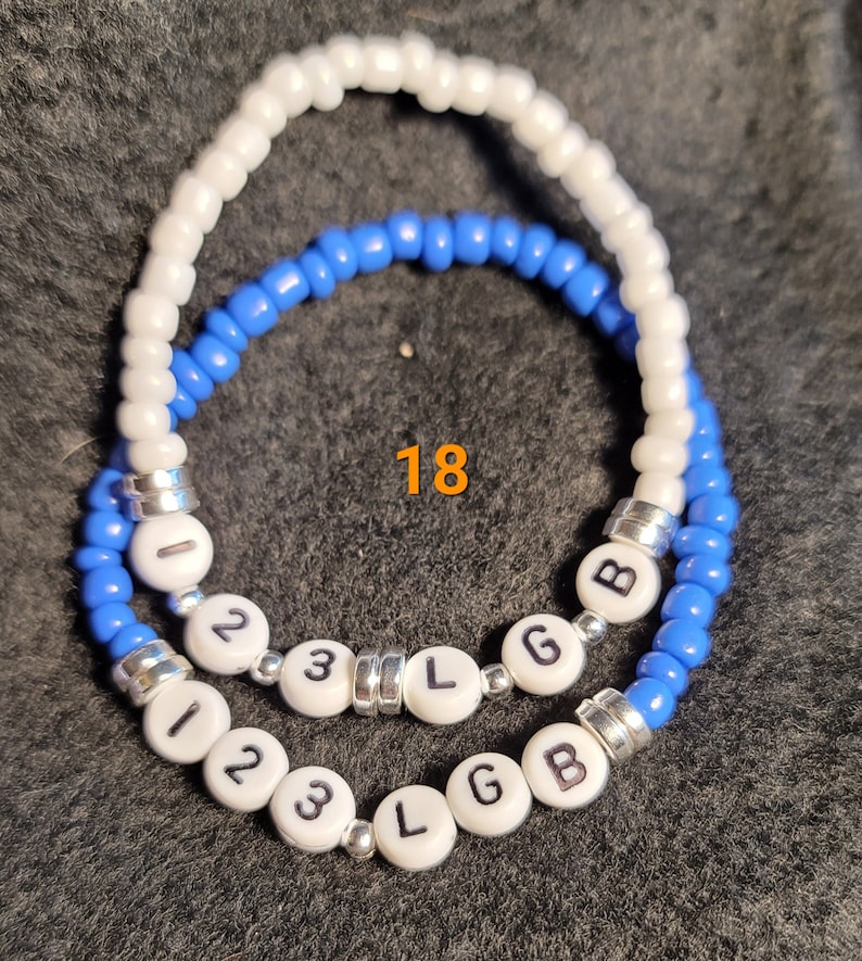 K&ouml;nnte beinhalten: Zwei Perlenarmb&auml;nder mit wei&szlig;en und blauen Perlen. Die Armb&auml;nder haben wei&szlig;e Perlen mit den Buchstaben "123 L G B" darauf. Die Zahl "18" ist auf dem Bild geschrieben.