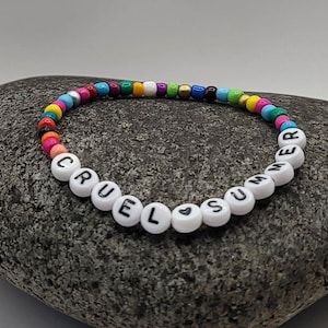 K&ouml;nnte beinhalten: Ein buntes Perlenarmband mit wei&szlig;en Buchstabenperlen, die "Cruel Summer" buchstabieren, mit einem kleinen Herzsymbol zwischen den W&ouml;rtern.