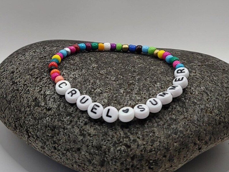 K&ouml;nnte beinhalten: Ein buntes Perlenarmband mit wei&szlig;en Buchstabenperlen, die "Cruel Summer" buchstabieren, mit einem kleinen Herzsymbol zwischen den W&ouml;rtern.