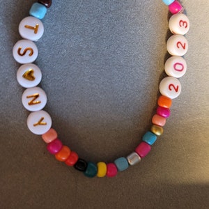K&ouml;nnte beinhalten: Ein farbenfrohes Perlenarmband mit wei&szlig;en Perlen, die "SONY" buchstabieren, mit einem goldenen Herzen zwischen dem "O" und dem "N" und wei&szlig;en Perlen, die "2023" in Rot buchstabieren.