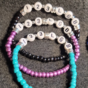 K&ouml;nnte beinhalten: Drei Perlenarmb&auml;nder mit den Buchstaben "123 LGB" auf den wei&szlig;en Perlen. Die Armb&auml;nder sind t&uuml;rkis, lila und schwarz.