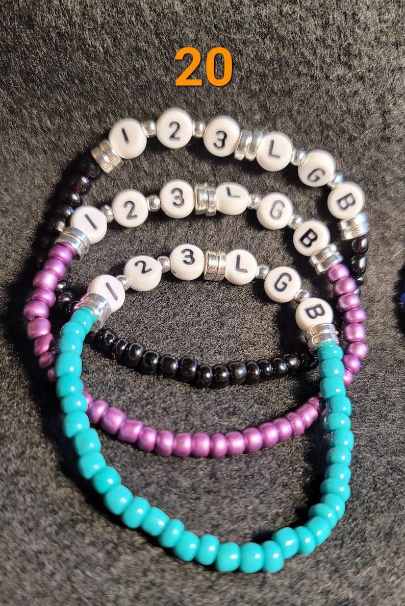 K&ouml;nnte beinhalten: Drei Perlenarmb&auml;nder mit den Buchstaben "123 LGB" auf den wei&szlig;en Perlen. Die Armb&auml;nder sind t&uuml;rkis, lila und schwarz.