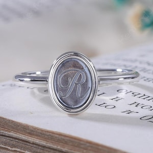 Könnte beinhalten: Ein silberfarbener Ring mit einer ovalen Vorderseite und einer polierten metallischen Oberfläche. In die Mitte des Ovals ist ein kursiver Buchstabe R eingraviert. Das Ringband ist dünn und kreisförmig und weist ein einheitliches Finish auf.