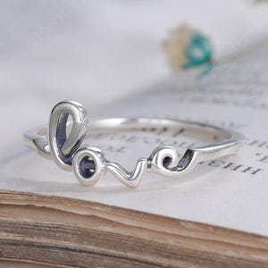 Könnte beinhalten: Ein silberner Ring mit dem Wort love in Schreibschrift. Die Buchstaben sind aus einem durchgehenden, polierten Silberband geformt und bilden ein filigranes, offenes Design, das auf den Seiten eines aufgeschlagenen Buches liegt.