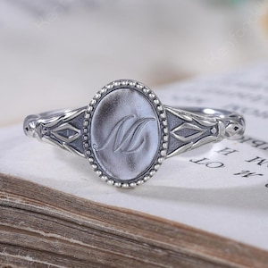 Op de afbeelding: Een zilveren ring met een ovaalvormig middendeel met een kralenrand en een gegraveerde letter M. De ringband heeft aan beide zijden ingewikkelde geometrische details en toont een gepolijste metalen afwerking met donkere accenten in de verdiepte delen.