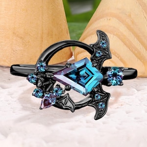 Może przedstawiać: Czarny pierścionek z kwadratowym, jasnoniebieskim kamieniem szlachetnym pośrodku, otoczonym mniejszymi niebieskimi i fioletowymi kamieniami. Pierścionek posiada motywy w kształcie nietoperzy po obu stronach centralnego kamienia, na białej powierzchni.