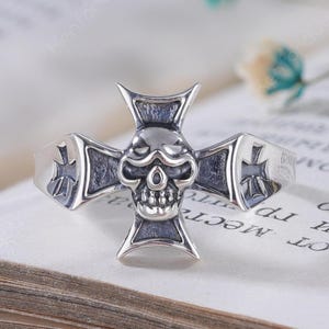 Könnte beinhalten: Ein silberner Ring mit einem zentralen Totenkopfmotiv, das in eine Kreuzform integriert ist. Das Kreuzdesign wird durch kleinere Kreuzgravuren auf der Ringschiene akzentuiert. Das Metall hat ein poliertes Finish mit abgedunkelten Vertiefungen.