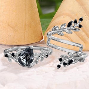 Könnte beinhalten: Drei silberne Ringe mit Blatt- und schwarzen Steinakzenten. Ein Ring hat einen großen, birnenförmigen, dunkelgrauen Stein. Die anderen beiden Ringe haben ein Wickeldesign mit schwarzen Steinen. Die Ringe werden auf einer weißen Oberfläche mit Holzkegeln im Hintergrund präsentiert.