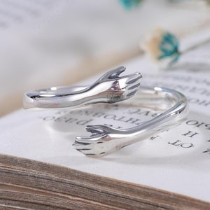 Könnte beinhalten: Ein offener Ring in polierter Silberoptik mit einem Design aus zwei Händen, die sich in einer Umarmung entgegenstrecken. Die Ringschiene ist glatt und reflektierend, wobei sich die Handmotive in der Mitte der verstellbaren Öffnung treffen.