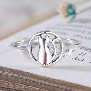 May include: A silver-toned ring featuring a circular frame with a stylized cat silhouette in the center. The cat is depicted in a seated position with its tail curved along the inner edge of the circular band, creating a polished metallic finish.
