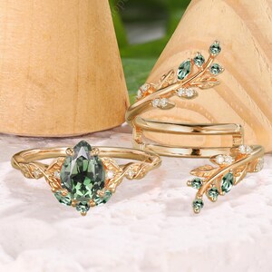 Könnte beinhalten: Eine Sammlung goldener Ringe mit grünen Edelsteinen und blattartigen Designs. Ein Ring hat einen birnenförmigen grünen Stein, während andere kleinere grüne Akzente aufweisen. Die Ringe werden auf einer weißen Oberfläche mit kegelförmigen Holzständern präsentiert.
