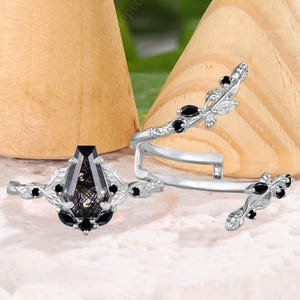 Könnte beinhalten: Ein silbernes Ring-Set mit einem zentralen Ring mit einem schwarzen Edelstein und kleineren schwarzen Steinen, begleitet von einem passenden Wickelring und einem kleineren Ring mit schwarzen Akzenten. Die Ringe werden auf einer weißen Oberfläche präsentiert.