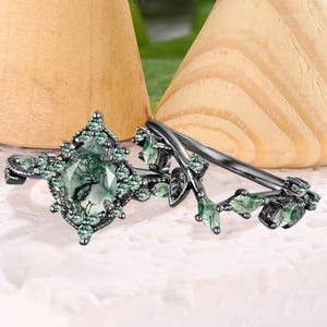 Könnte beinhalten: Zwei schwarze Ringe mit grünen Edelsteinen. Ein Ring hat einen großen, ovalen grünen Stein, der von kleineren grünen Steinen umgeben ist. Der andere Ring hat ein Blumendesign mit grünen Akzenten. Die Ringe werden auf einer weißen Oberfläche präsentiert.