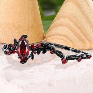 May include: A black ring set with a marquise-cut, deep red gemstone and smaller red stones. The ring features a leaf and vine design. The set includes a matching band with red accents. The rings are displayed on a white surface with wooden cones in the background.