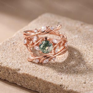 May include: A rose gold ring featuring a round moss agate centre stone surrounded by a branch-like band design. The band is adorned with small, clear, marquise and round-cut stones set along the metal leaves and vines encircling the centre.