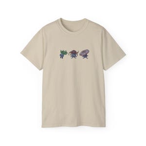 Peut inclure: Un t-shirt beige avec un imprimé graphique de trois personnages de dessin animé. Les personnages sont bleus, verts et violets et ont des formes de tête différentes.