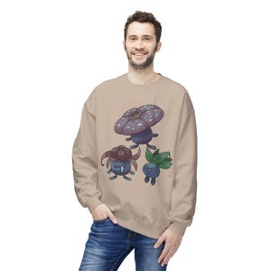 Peut inclure: Un sweat-shirt beige avec un motif coloré de trois personnages de champignons de dessin animé. Le sweat-shirt a un col rond et est fait d'une matière douce. Le motif comprend un grand champignon violet et deux petits personnages de champignons bleus.