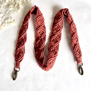 Puede incluir: Una larga correa de macramé marrón con dos cierres metálicos. La correa está hecha de un cordón grueso y trenzado y tiene un patrón decorativo.