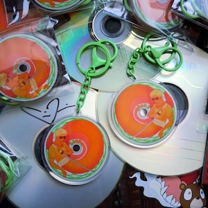 Può includere: Una collezione di CD e portachiavi. I portachiavi presentano un design circolare con sfondo arancione e bianco e un personaggio dei cartoni animati. I portachiavi hanno chiusure e catene verdi. I CD sono argentati.