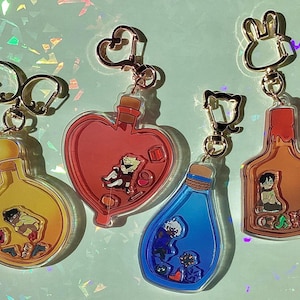 May include: Four colorful keychains featuring glass bottles with different characters inside. The bottles are filled with red, blue, and orange liquids. Each keychain has a gold metal ring and a different style of clasp.