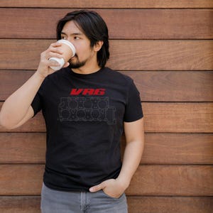 Può includere: Una t-shirt nera a maniche corte con un logo V6 rosso sopra un'illustrazione a linee bianche di un blocco motore V6. Il capo presenta un girocollo e una vestibilità standard, con un design grafico centrato sulla parte del petto.