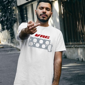 Può includere: Una t-shirt bianca a maniche corte con una stampa grafica che raffigura il contorno grigio di una guarnizione della testata del motore con il testo rosso VR6 sopra. Il design è centrato sul petto del capo, che presenta un girocollo.