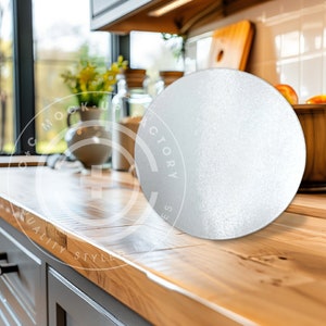 May include: A round, silver cake board sits on a wooden countertop in a kitchen. The board is blank and ready for a cake to be placed on top.