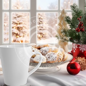 May include: A white ceramic mug sits on a white tablecloth next to a glass serving dish filled with powdered sugar-dusted cookies. A small Christmas tree, red ornament, and pinecone are also on the table, with a snowy window in the background.