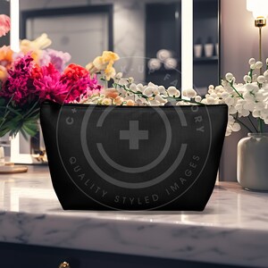 May include: Black zippered makeup bag with a white circle and a plus sign in the center. The text "Quality Styled Images" is printed around the circle. The bag is sitting on a white countertop.