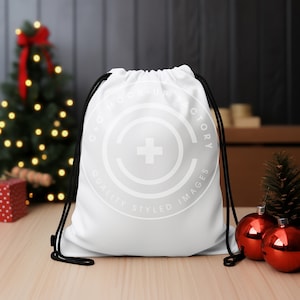 Puede incluir: Una bolsa blanca de cordón con cordones negros. La bolsa tiene un logotipo circular blanco con una cruz en el centro y el texto "GOX MOCK UP FACTORY QUALITY STYLED IMAGES". La bolsa está sobre una mesa de madera con un árbol de Navidad y adornos rojos en el fondo.