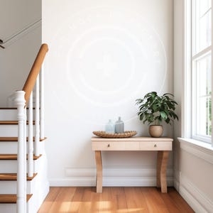 Puede incluir: Una consola de madera clara con dos cajones, que sostiene una bandeja tejida con dos botellas de vidrio y una pequeña planta en maceta. La mesa está colocada contra una pared blanca con una escalera y una ventana en el fondo.