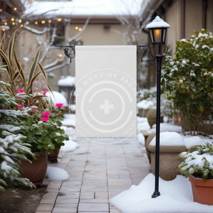 May include: A white blank sign with black metal brackets hangs from a black metal pole in a snowy winter scene. The sign is in front of a brick building with a lamppost and snow-covered plants.